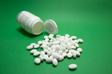 Viele weiße Tabletten liegen auf einer grünen Oberfläche neben einer geöffneten Medikamentendose.