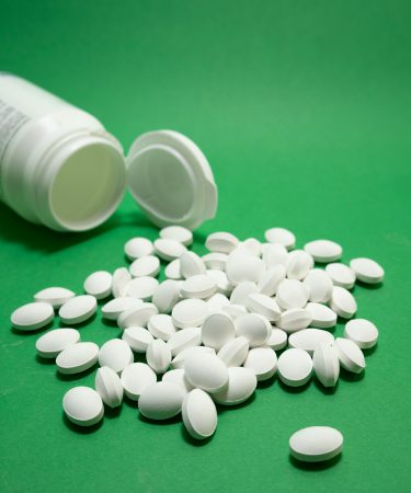 Viele weiße Tabletten liegen auf einer grünen Oberfläche neben einer geöffneten Medikamentendose.