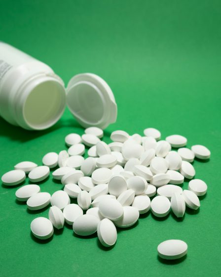 Viele weiße Tabletten liegen auf einer grünen Oberfläche neben einer geöffneten Medikamentendose.