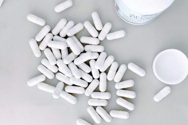 Zahlreiche weiße Kapseln liegen auf einer hellen Fläche neben einem geöffneten Behälter für Nahrungsergänzungsmittel.