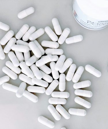 Zahlreiche weiße Kapseln liegen auf einer hellen Fläche neben einem geöffneten Behälter für Nahrungsergänzungsmittel.