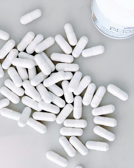 Zahlreiche weiße Kapseln liegen auf einer hellen Fläche neben einem geöffneten Behälter für Nahrungsergänzungsmittel.