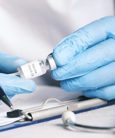 Eine medizinische Fachkraft mit blauen Handschuhen hält eine kleine Flasche mit der Aufschrift „Vaccine Covid-19“ neben medizinischen Instrumenten.