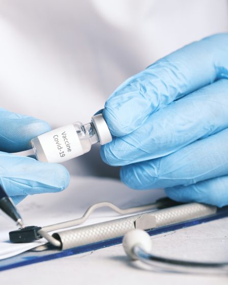 Eine medizinische Fachkraft mit blauen Handschuhen hält eine kleine Flasche mit der Aufschrift „Vaccine Covid-19“ neben medizinischen Instrumenten.