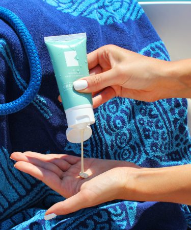 Eine Frau dosiert eine cremefarbene After-Sun-Lotion mit NL-GHK-Cu aus einer türkisfarbenen Tube in ihre Handfläche.