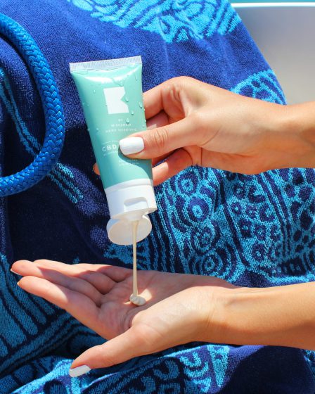 Eine Frau dosiert eine cremefarbene After-Sun-Lotion mit NL-GHK-Cu aus einer türkisfarbenen Tube in ihre Handfläche.