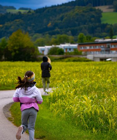 Ein junges Mädchen läuft im Sommer in einer rosafarbenen Jacke auf einem asphaltierten Weg entlang eines grünen Feldes.