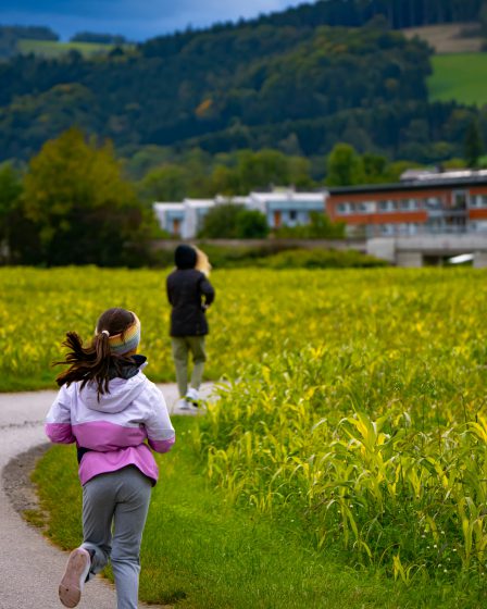 Ein junges Mädchen läuft im Sommer in einer rosafarbenen Jacke auf einem asphaltierten Weg entlang eines grünen Feldes.