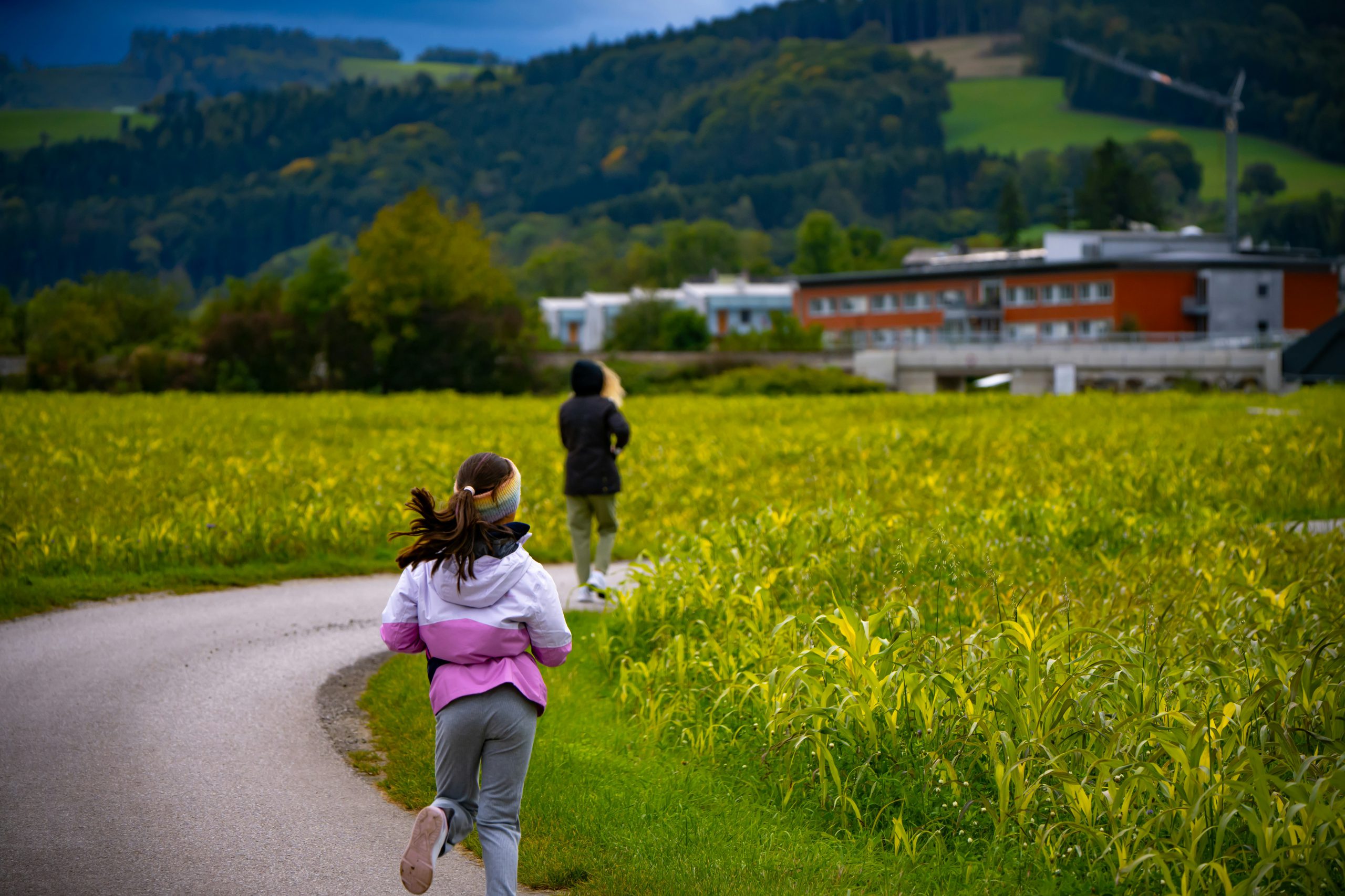 Ein junges Mädchen läuft im Sommer in einer rosafarbenen Jacke auf einem asphaltierten Weg entlang eines grünen Feldes.