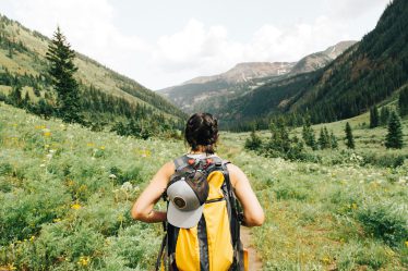 Frau mit gelbem Trekkingrucksack auf einem Bergpfad blickt in ein grünes Tal – aktive Regeneration nach einer Outdoor-Verletzung mit NL-BPC-157.