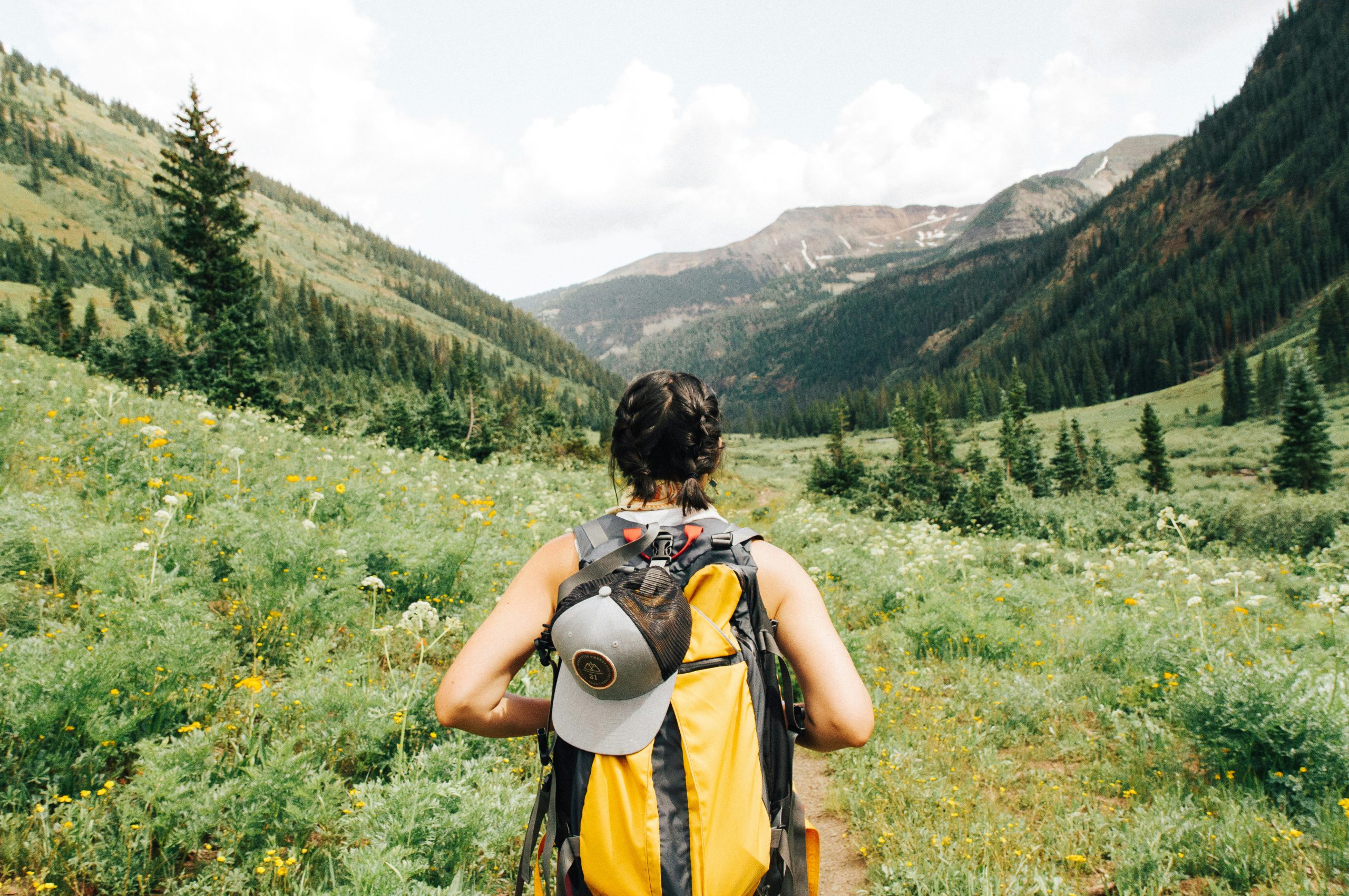 Frau mit gelbem Trekkingrucksack auf einem Bergpfad blickt in ein grünes Tal – aktive Regeneration nach einer Outdoor-Verletzung mit NL-BPC-157.